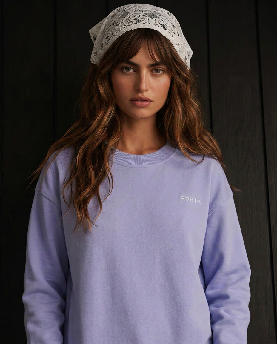 A woman wearing a light lavender crewneck sweatshirt with a small white embroidered 'POETA' logo on the chest, paired with a white lace headscarf, set against a dark wood-paneled background.