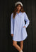 A woman modeling a light blue long-sleeve button-up shirt dress with chest pockets, paired with a white lace headband and beige socks, standing against a dark wood-paneled background.