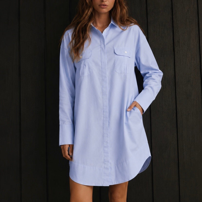 A woman modeling a light blue long-sleeve button-up shirt dress with chest pockets, paired with a white lace headband and beige socks, standing against a dark wood-paneled background.