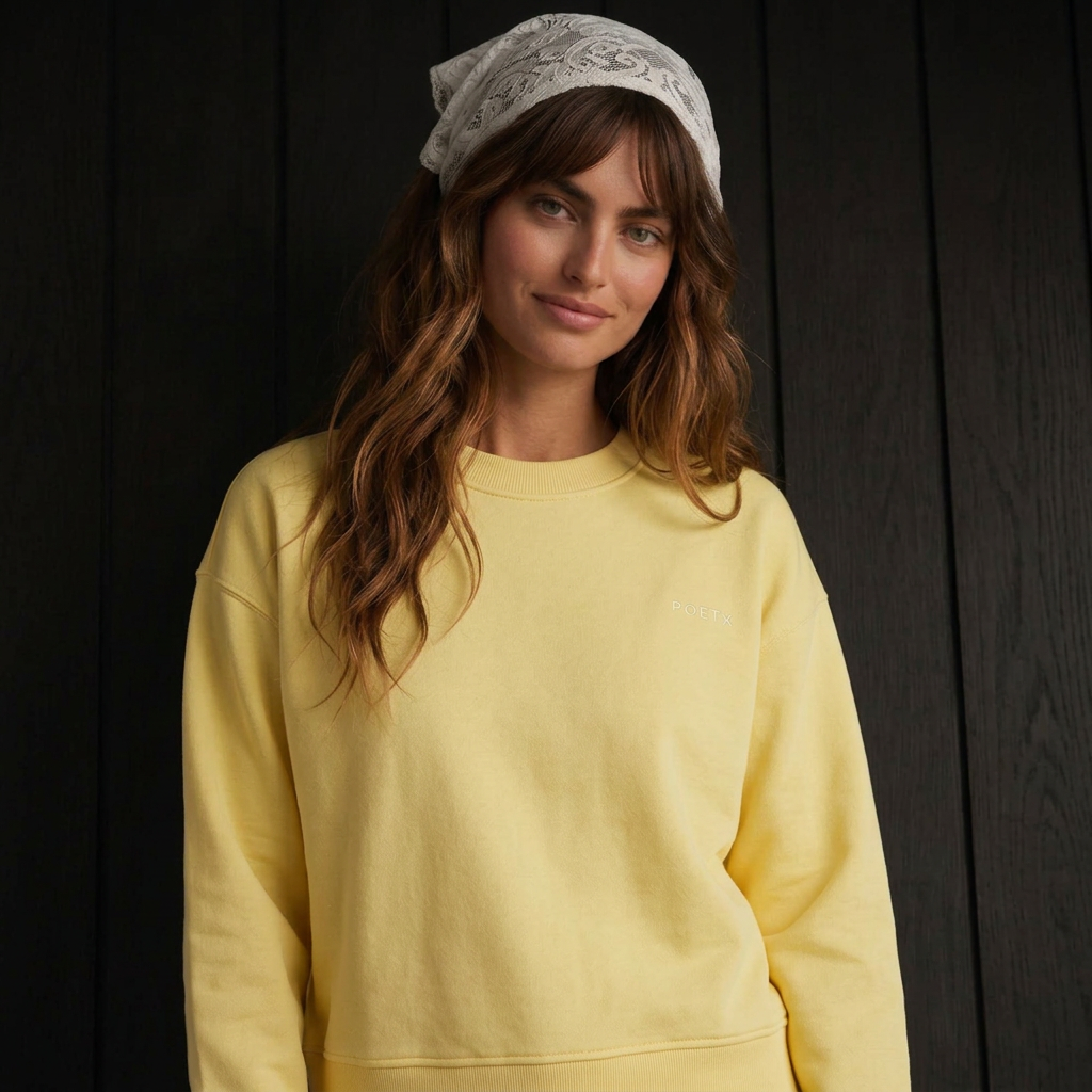 A woman wearing a pale yellow crewneck sweatshirt and a light gray patterned bandana, standing against a dark wood-paneled background.