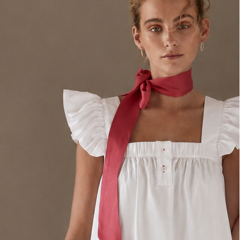 A model stands against a minimalist studio backdrop wearing a white ruffled short-sleeve top with a red neck scarf, matching white shorts, and white socks.