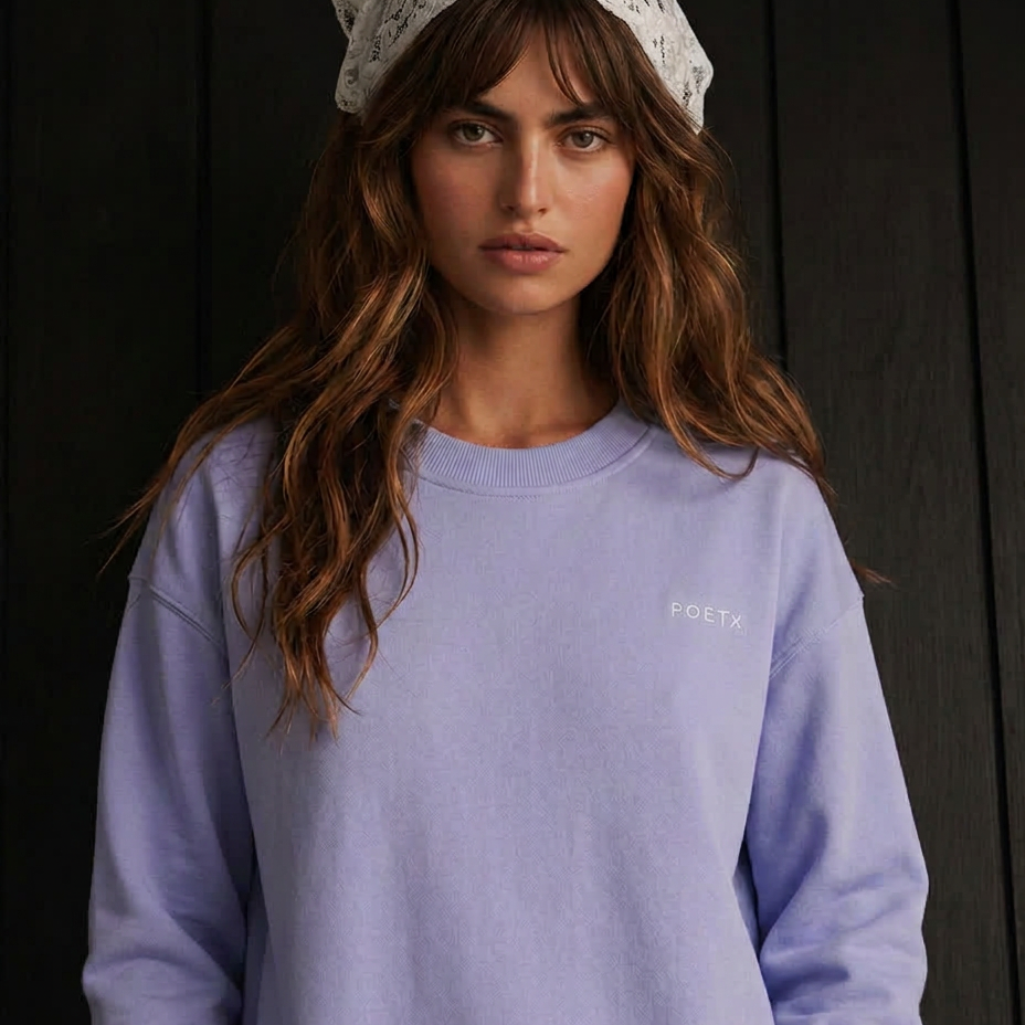 A woman wearing a light lavender crewneck sweatshirt with a small white embroidered 'POETA' logo on the chest, paired with a white lace headscarf, set against a dark wood-paneled background.