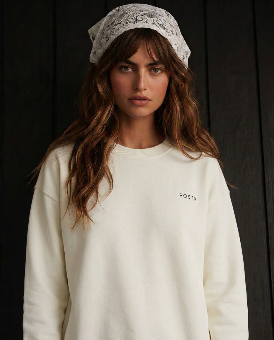 A woman wearing a cream-colored crewneck sweatshirt with a small black 'POETX' logo on the left chest, paired with a white patterned headscarf, set against a dark wood-paneled background.