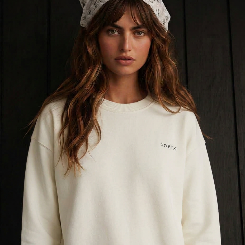 A woman wearing a cream-colored crewneck sweatshirt with a small black 'POETX' logo on the left chest, paired with a white patterned headscarf, set against a dark wood-paneled background.